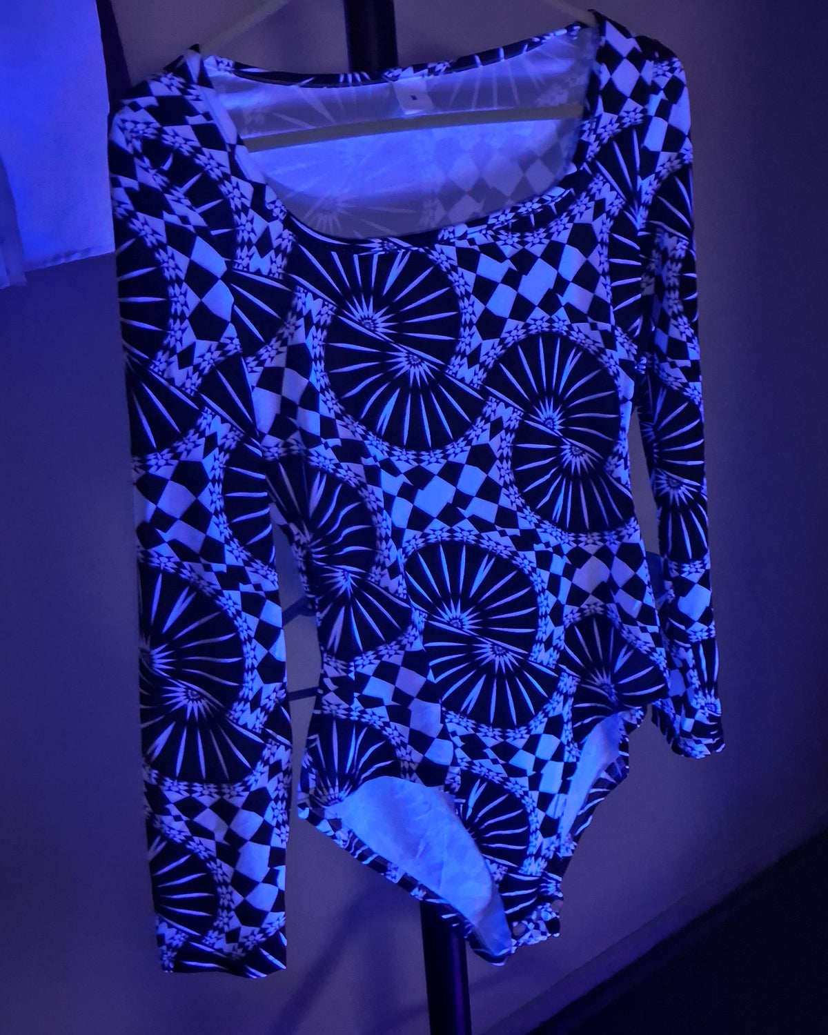 A long-sleeve bodysuit with a black and blue geometric pattern featuring circular fan and checkerboard motifs, hanging on a black hanger against a dark background under blue light.